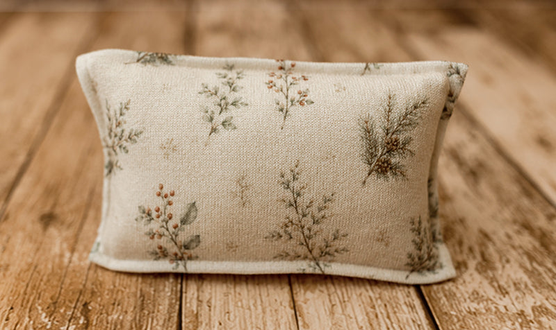 Mantle Garland newborn photography pillowcase featuring delicate evergreen branches, sage green botanical sprigs, and subtle rust-colored berries on a warm cream textured fabric, displayed on a rustic wooden farmhouse backdrop.