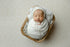 Newborn swaddled in ivory fuzzy textured lace fabric, resting in a wooden basket prop with a matching bonnet.