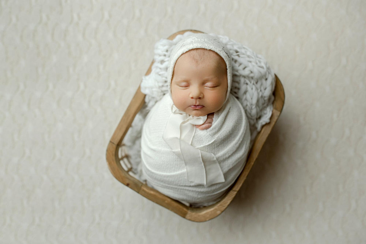 Newborn swaddled in ivory fuzzy textured lace fabric, resting in a wooden basket prop with a matching bonnet.