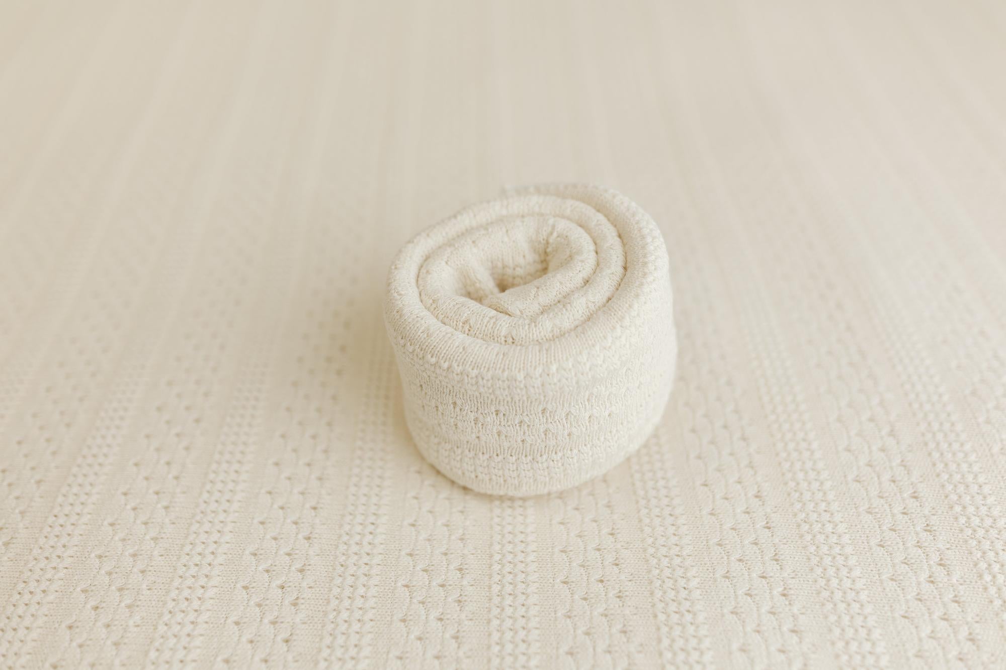 A rolled cream-colored knit blanket or fabric piece, shown on a matching textured cream background.