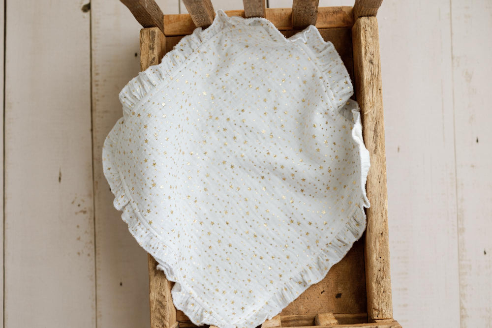A white eyelet muslin fabric with tiny gold sequins and a ruffled edge, draped inside a rustic wooden crate against a light wood plank background.