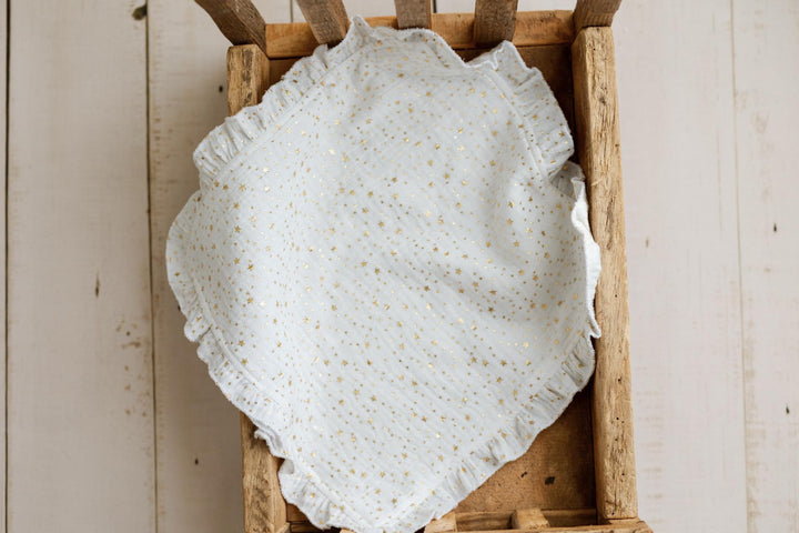 A white eyelet muslin fabric with tiny gold sequins and a ruffled edge, draped inside a rustic wooden crate against a light wood plank background.