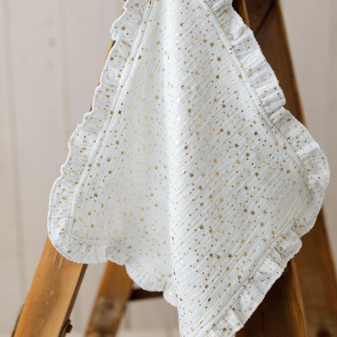 A white cotton muslin baby blanket with gold glitter speckles and ruffled edges, draped over a rustic wooden step ladder.