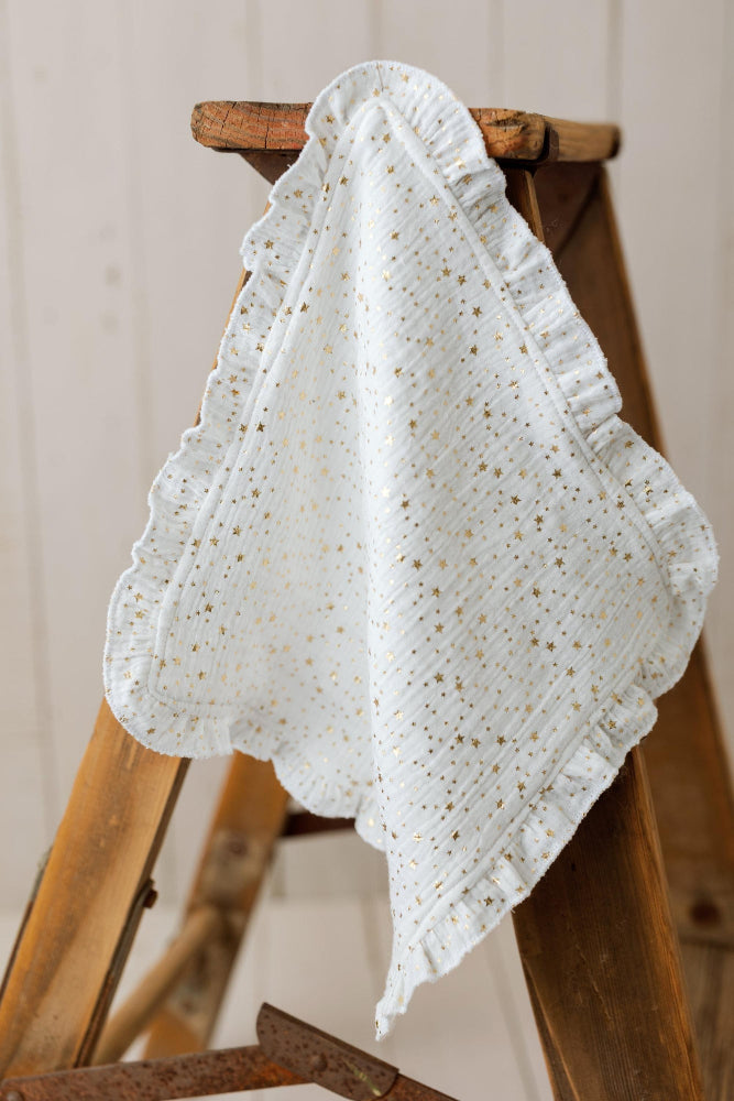 A white cotton muslin baby blanket with gold glitter speckles and ruffled edges, draped over a rustic wooden step ladder.