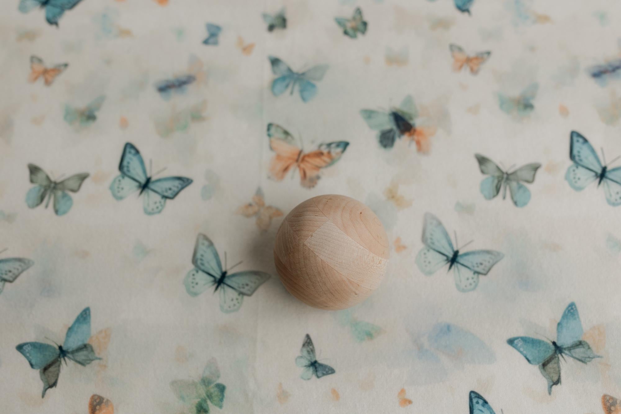 Creamy neutral newborn posing fabric with sage, teal, and peach butterfly prints and a smooth wooden sphere prop.