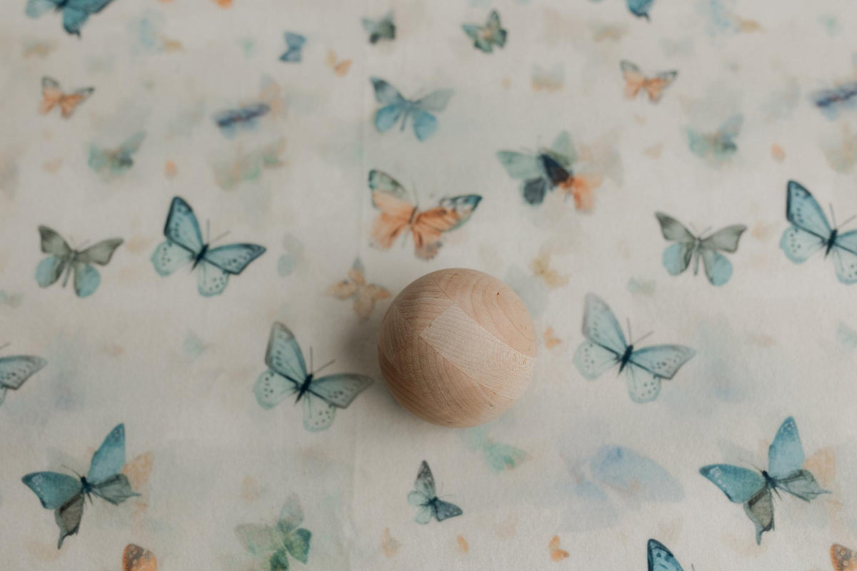 Creamy neutral newborn posing fabric with sage, teal, and peach butterfly prints and a smooth wooden sphere prop.