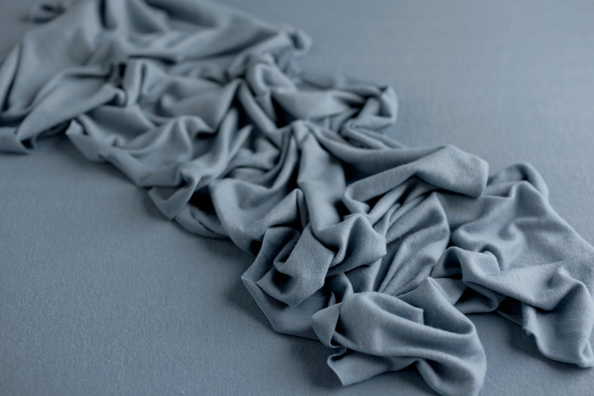 Deep dusty blue cashmere-style wrap featuring soft, draped fabric folds on a matching textured background.