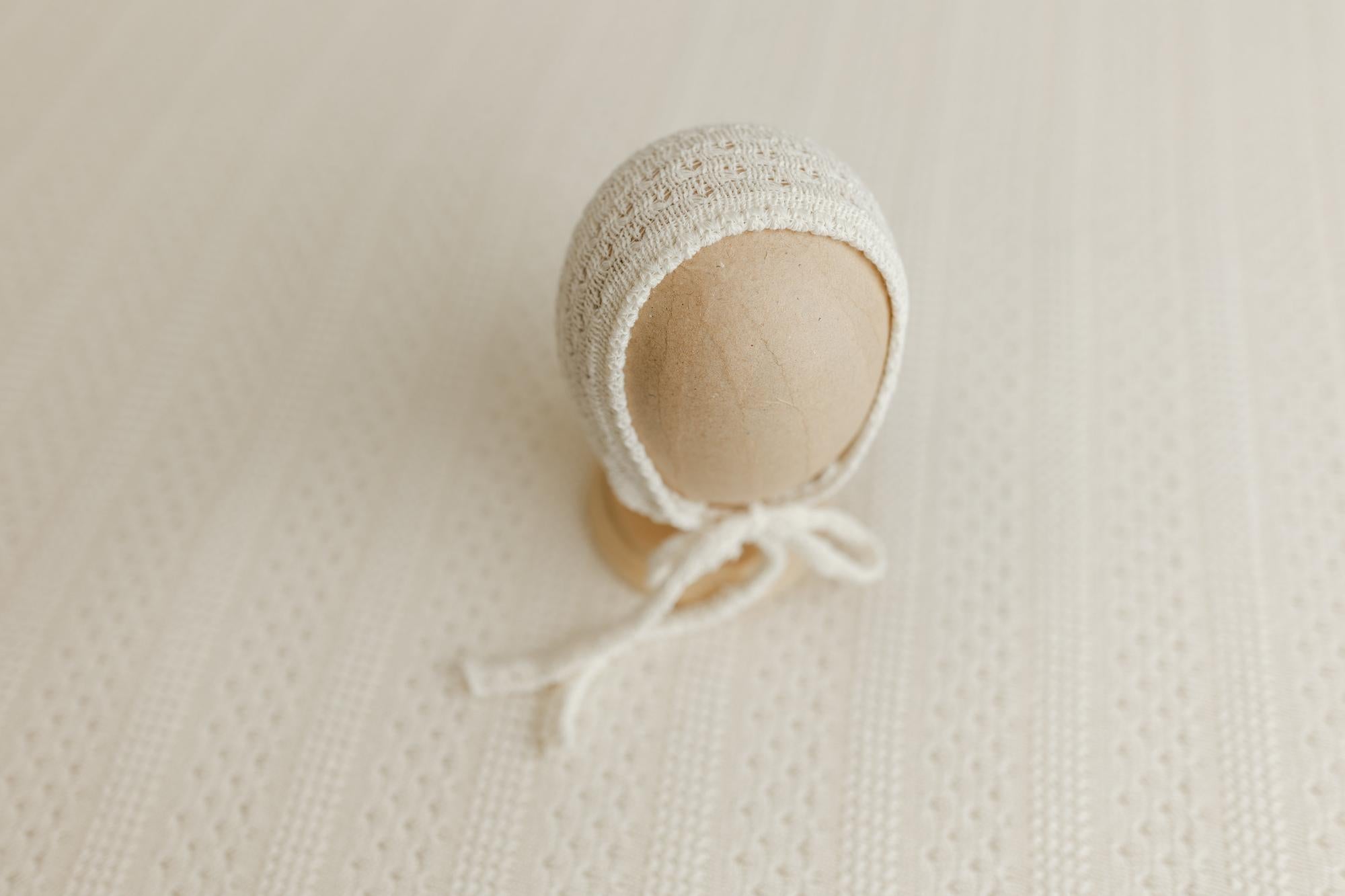 Cream knitted baby bonnet on a textured beige surface