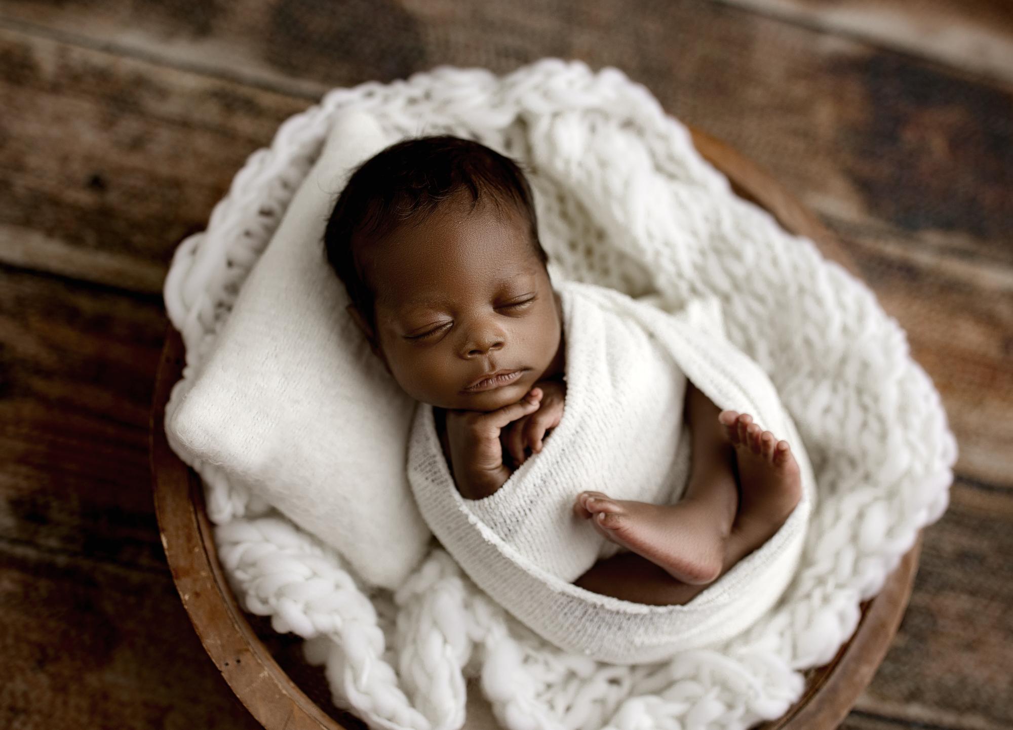 Newborn swaddled in a soft ivory Nest wrap, resting on a chunky knit fabric inside a wooden bowl prop.