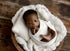 Newborn swaddled in a soft ivory Nest wrap, resting on a chunky knit fabric inside a wooden bowl prop.