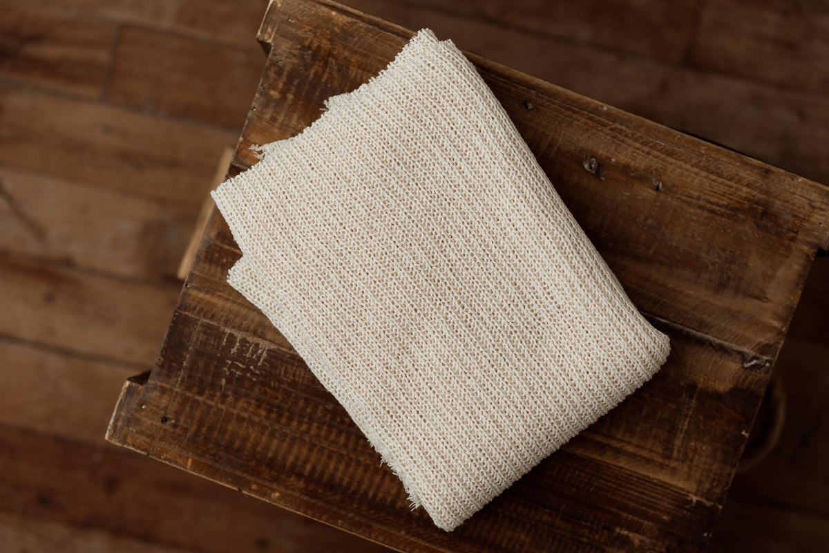 Beige, open-knit newborn photography fabric wrap with a textured, ramen-toned finish folded on a rustic wooden crate.