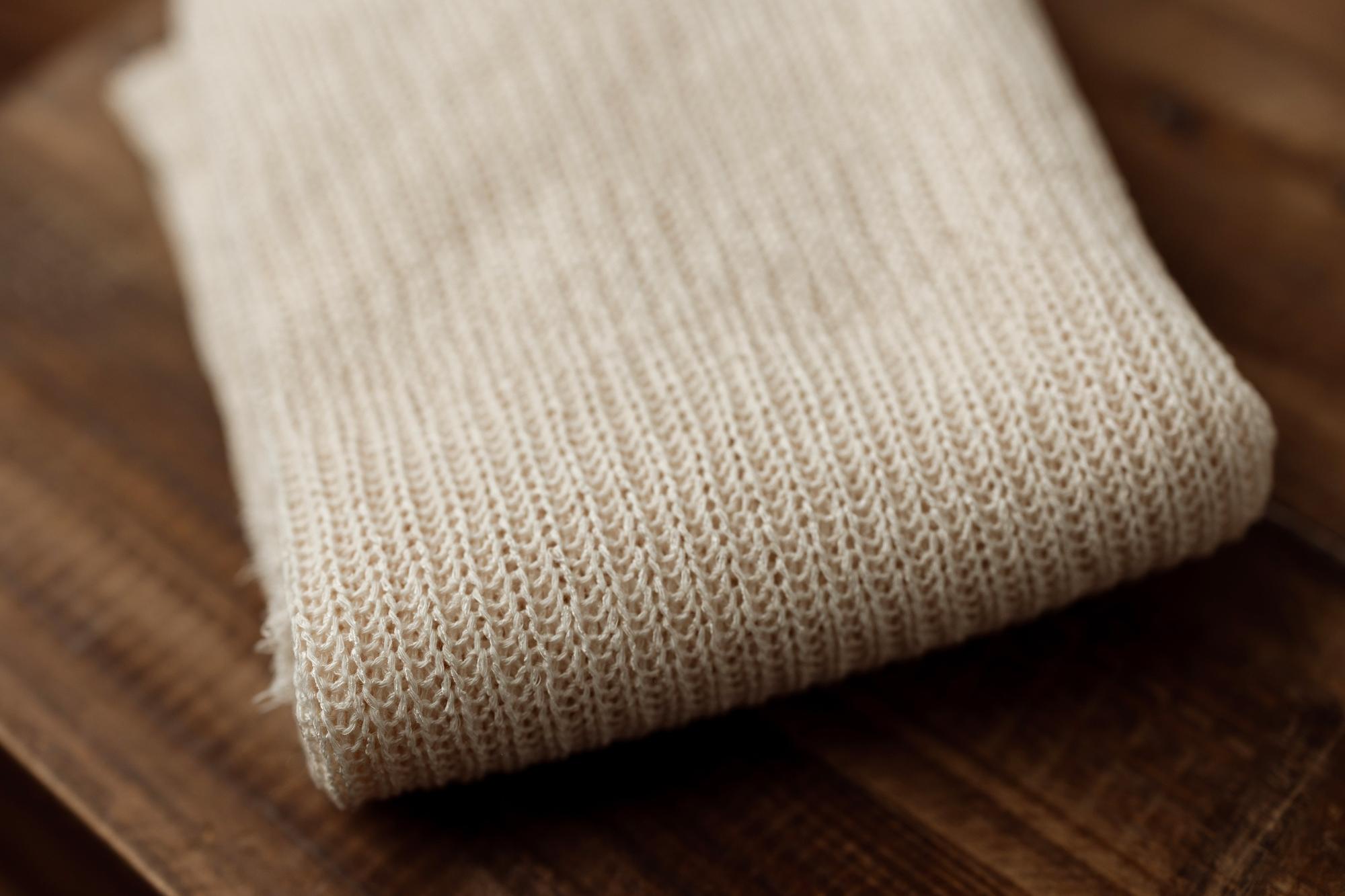 Folded beige ramen-toned newborn photography fabric with a soft, open-knit texture resting on a dark wooden surface.