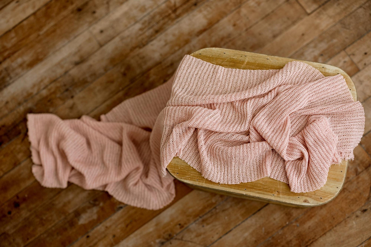 Soft strawberry milk pink open-knit newborn photography wrap with a textured, tattered edge draped over a wooden stool.