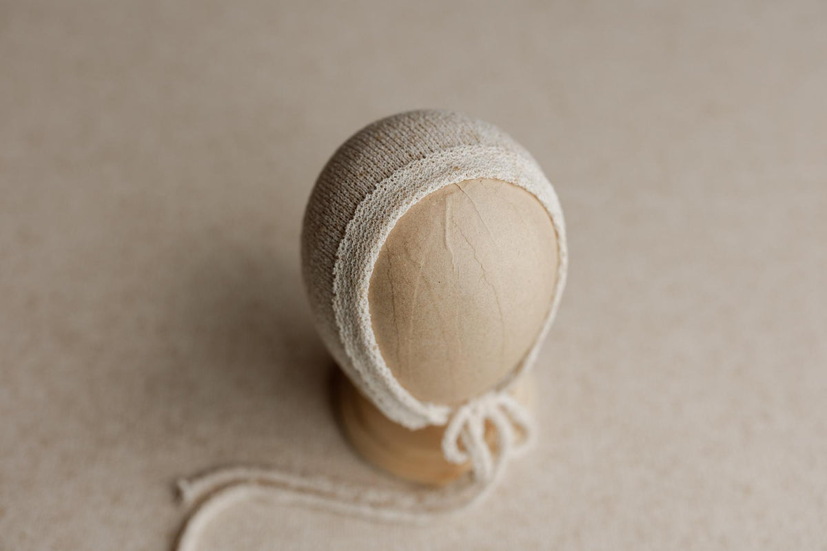 Neutral beige newborn photography bonnet with a delicate textured trim and tie strings on a mannequin head.