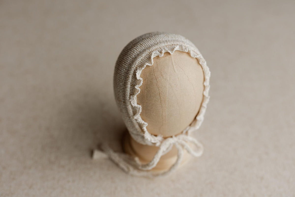 Neutral Seagrove knit newborn bonnet with delicate lace trim and tie closure on a wooden display head.
