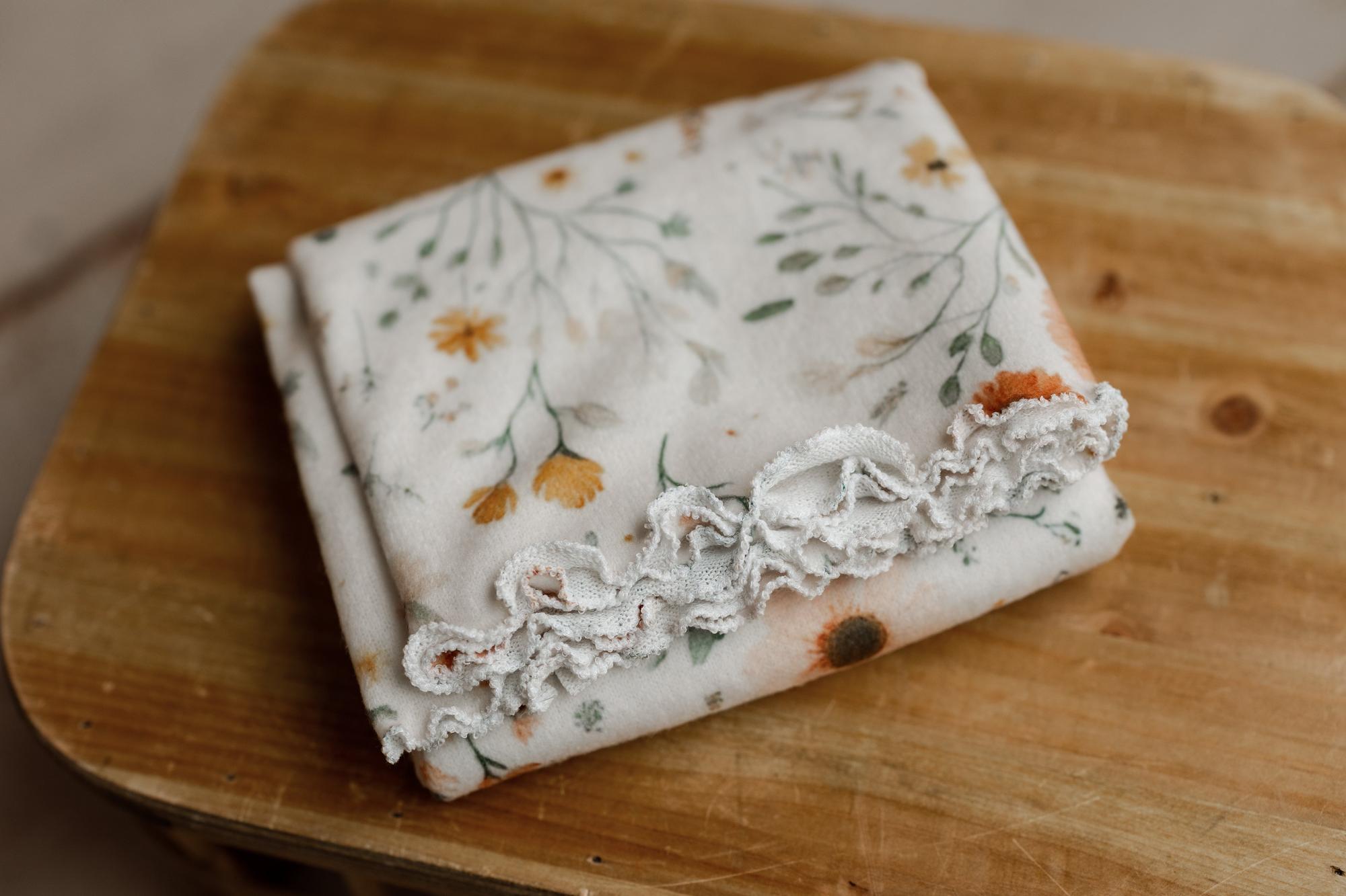 Folded newborn photography wrap with a peach, gold, and sage floral print and delicate white ruffled lace trim.