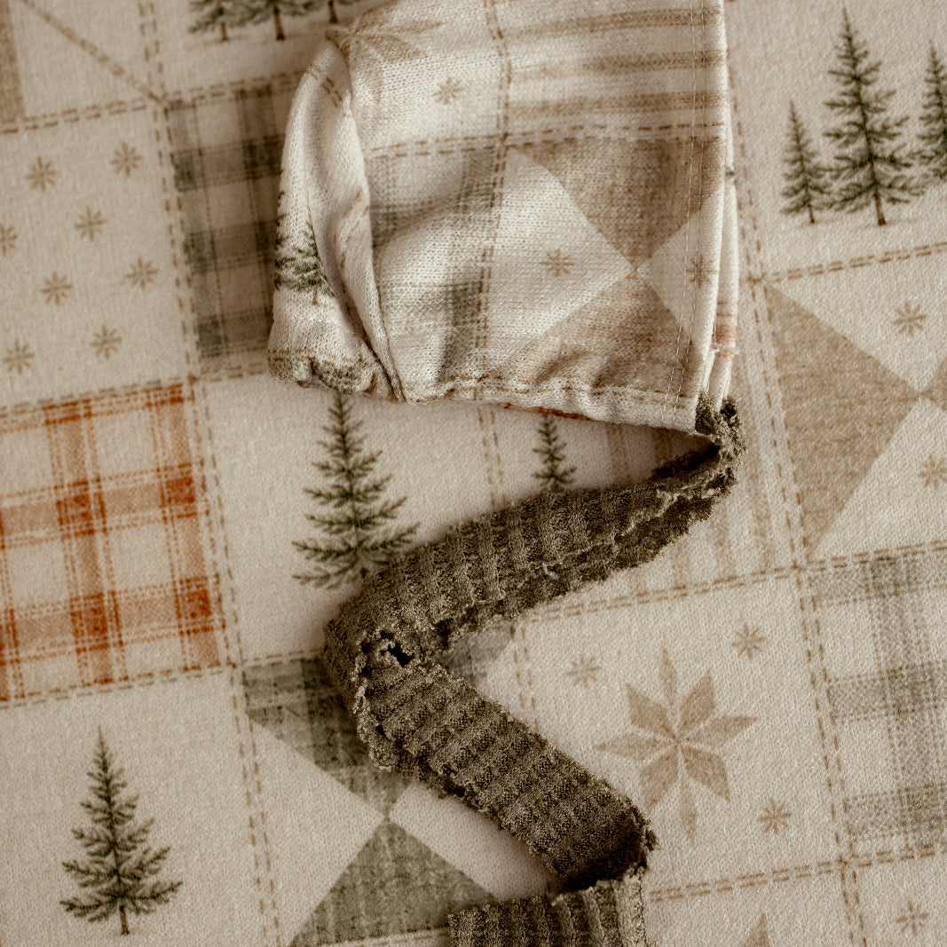 A soft fabric baby bonnet with a knotted tie strap, laid flat on a quilted patchwork fabric background featuring pine trees and geometric patterns.