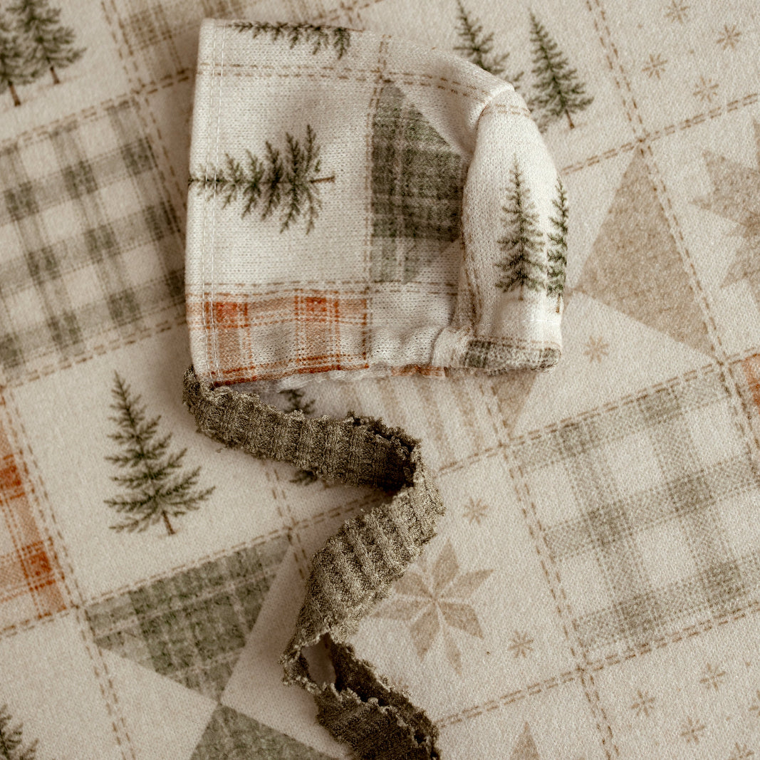 A folded cream-colored fabric item with a rustic pine tree and plaid pattern, tied with a matching knotted ribbon, laid on a quilted fabric surface.