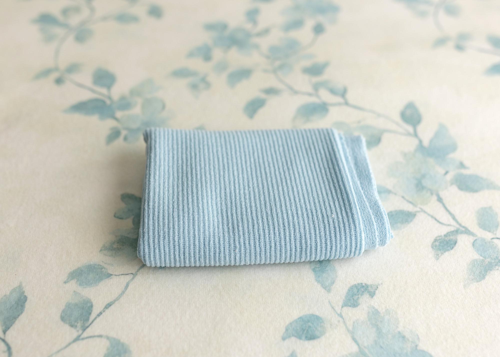 Folded blue ribbed newborn wrap resting on ivory fabric with a vintage watercolor blue floral vine print.