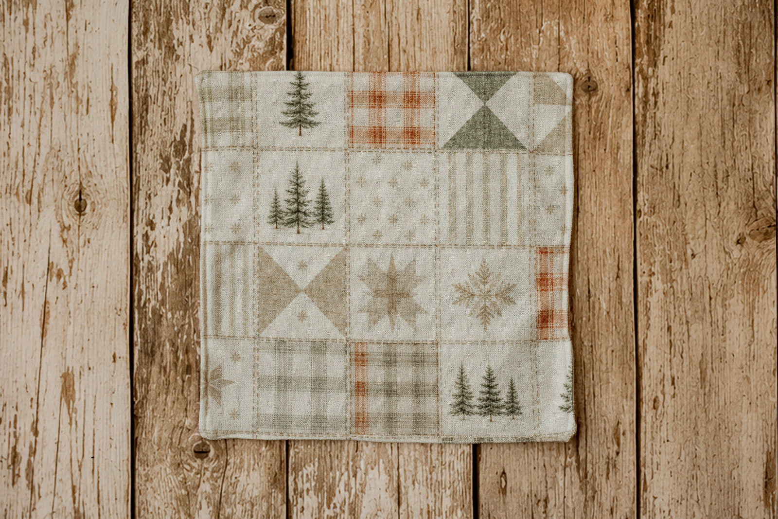A newborn photography mini blanket featuring a rustic patchwork design with pine trees, plaid, stripes, and snowflake motifs in muted earth tones, displayed flat on a weathered wooden plank background.
