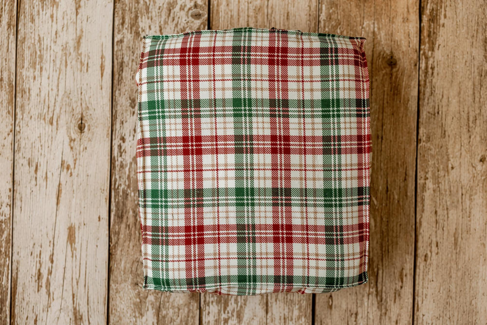 A square fabric cushion or pillow with a red, green, and white plaid pattern, photographed from above on a rustic light brown wooden plank surface.