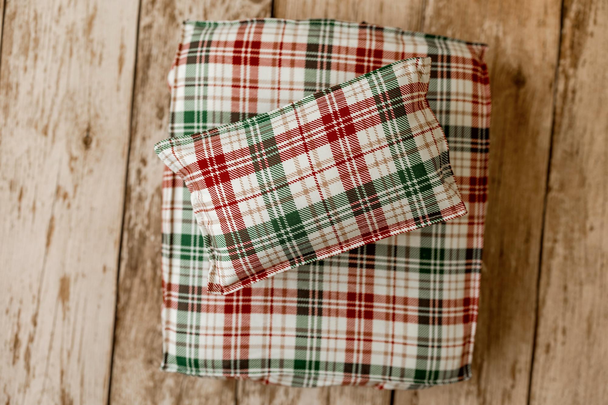 A small rectangular pillow with a red, green, and white plaid pattern rests on top of a larger matching pillow or cushion on a rustic wooden plank surface.