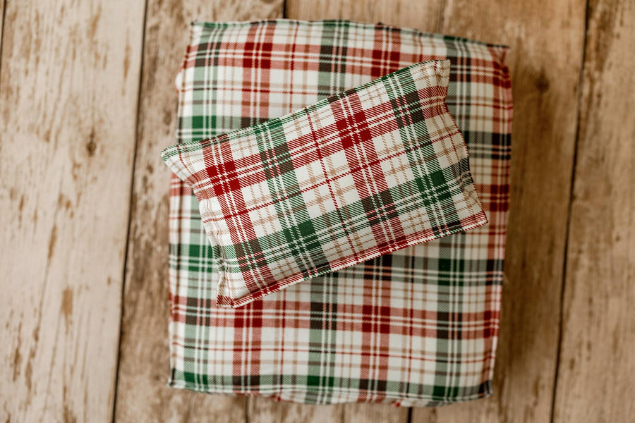 A small rectangular pillow with a red, green, and white plaid pattern rests on top of a larger matching pillow or cushion on a rustic wooden plank surface.