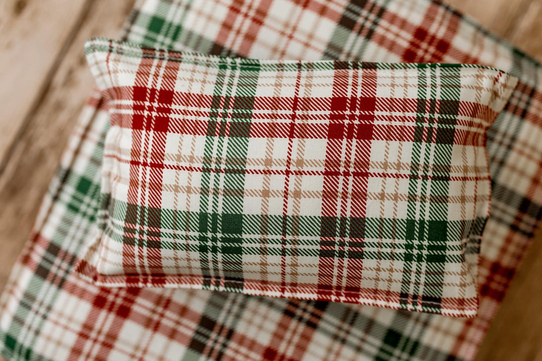 A small rectangular pillow with a red, green, and white plaid pattern, resting on a matching plaid blanket on a light wood surface.