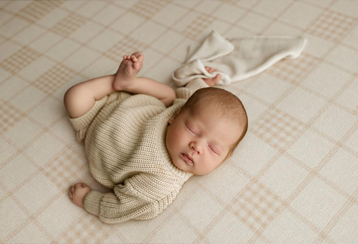 Sleeping newborn baby wearing a cream knit outfit posed on Grandpa's Twin beige plaid posing fabric for a soft, neutral newborn photography setup.