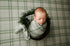 Newborn swaddled in sage green plaid fabric, resting in a rustic bucket with matching vintage-style posing wrap.