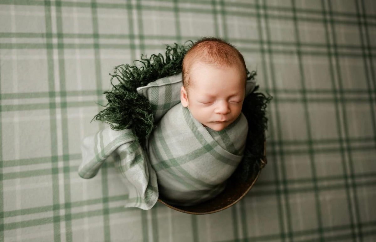 Newborn swaddled in sage green plaid fabric, resting in a rustic bucket with matching vintage-style posing wrap.