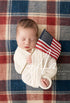 Newborn baby wrapped in cream knit fabric resting on a red, white, and blue plaid posing fabric with a small US flag.