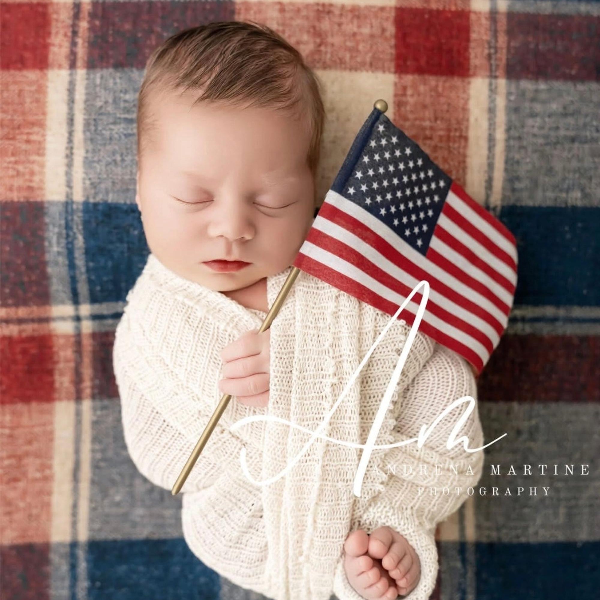 Newborn baby wrapped in cream knit fabric resting on a red, white, and blue plaid posing fabric with a small US flag.