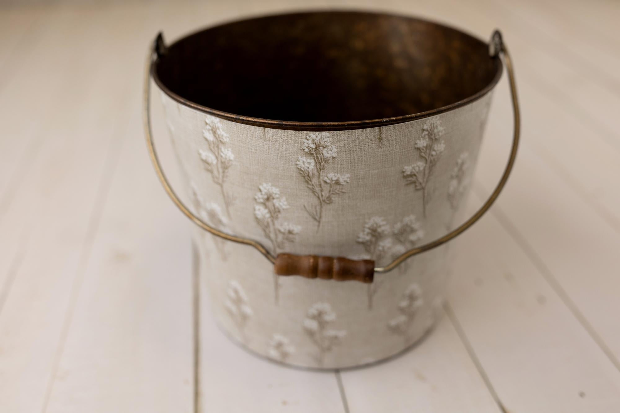 A vintage-style metal bucket with a distressed white floral pattern and a dark brown handle, set on a light-colored wooden plank surface.