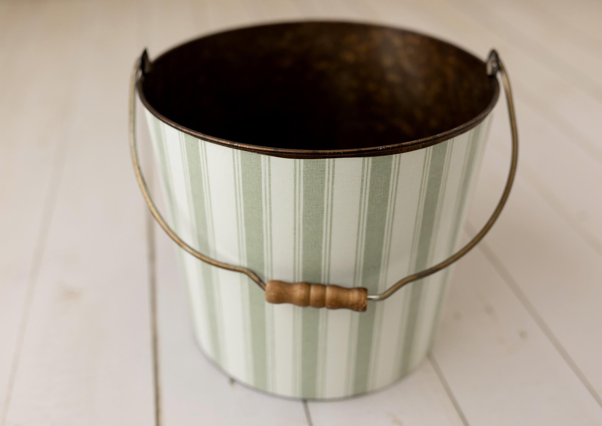 A vintage-style metal bucket with vertical sage green and white stripes, a dark bronze-toned interior, and a curved metal handle with a wooden grip, shown on a white wooden surface.
