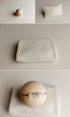 A soft cream-colored textured fabric accessory, shown folded and laid flat on a neutral fabric backdrop, with a small wooden ball and a pillow-like prop in the background.