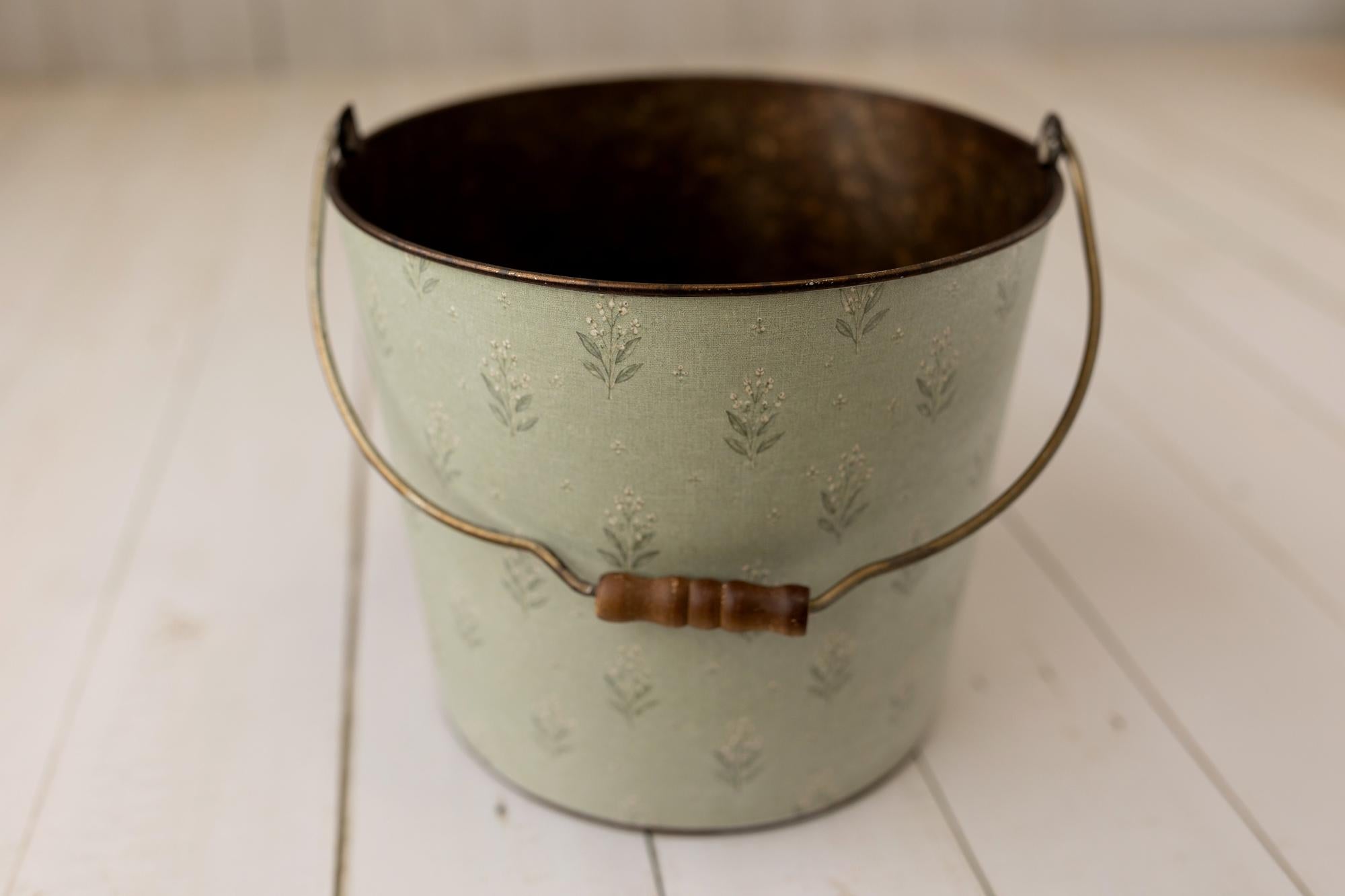 A vintage-style metal bucket with a pale sage green embossed floral pattern, a dark brown interior, and a curved wire handle with a brown wooden connector, set on a white wooden plank floor.