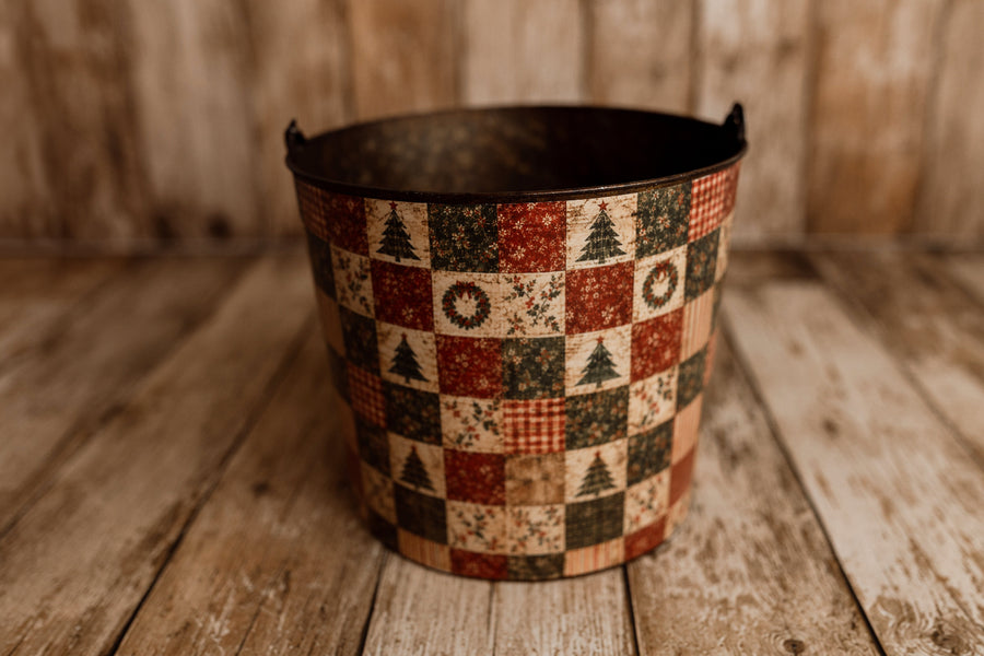 Very Merry Christmas bucket cover displayed on a rustic metal newborn posing bucket, featuring vintage red and green holiday patchwork with trees, wreaths, and plaid.
