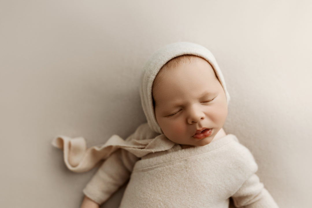 Newborn in sand knit romper with matching bonnet posed on neutral fabric backdrop