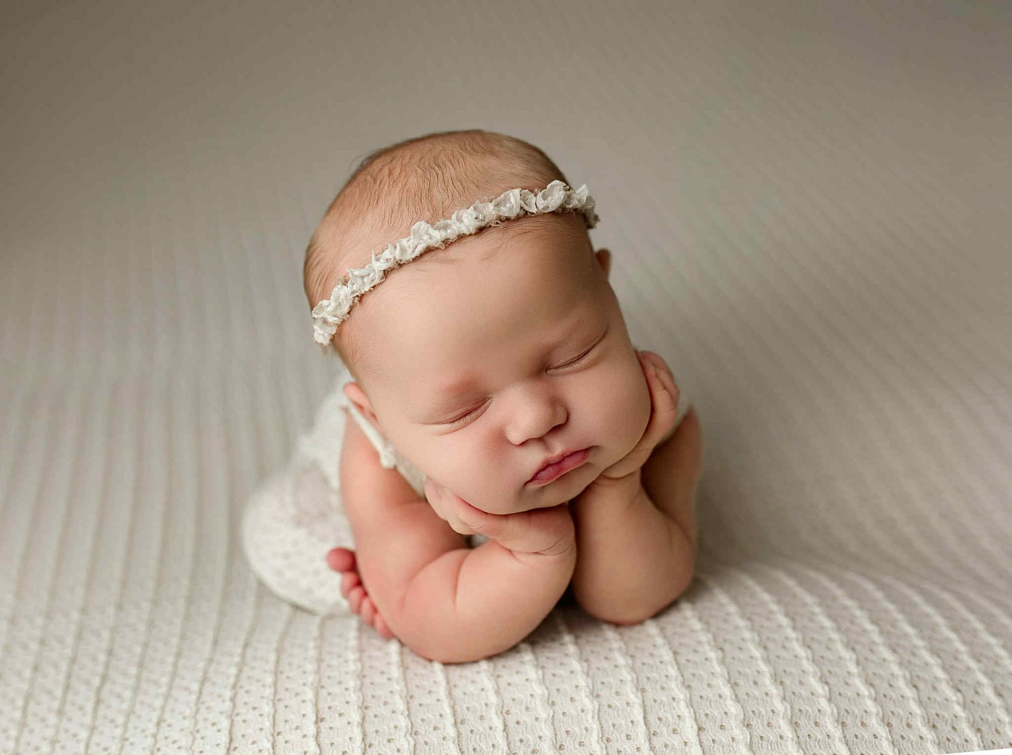 Newborn baby sleeping on off-white textured knit posing fabric with matching wrap and delicate floral headband.