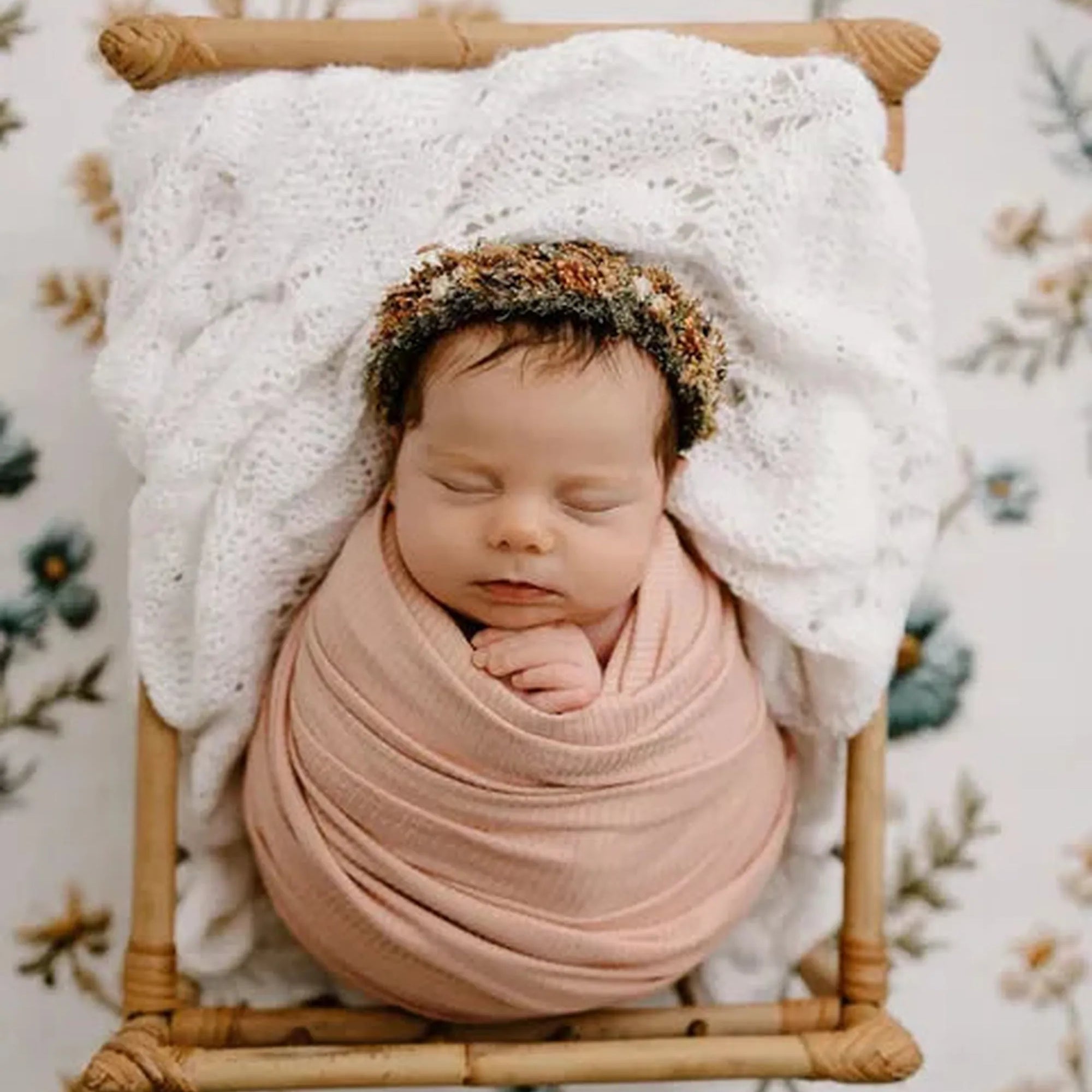 A sleeping newborn baby swaddled in a soft dusty pink blanket and wearing a floral headband, resting inside a small woven rattan basket cradled by a white knit blanket against a floral patterned backdrop.