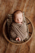 Newborn baby wrapped in a coffee brown knit blanket, resting on brown plaid sheets and a matching pillow in a round basket.