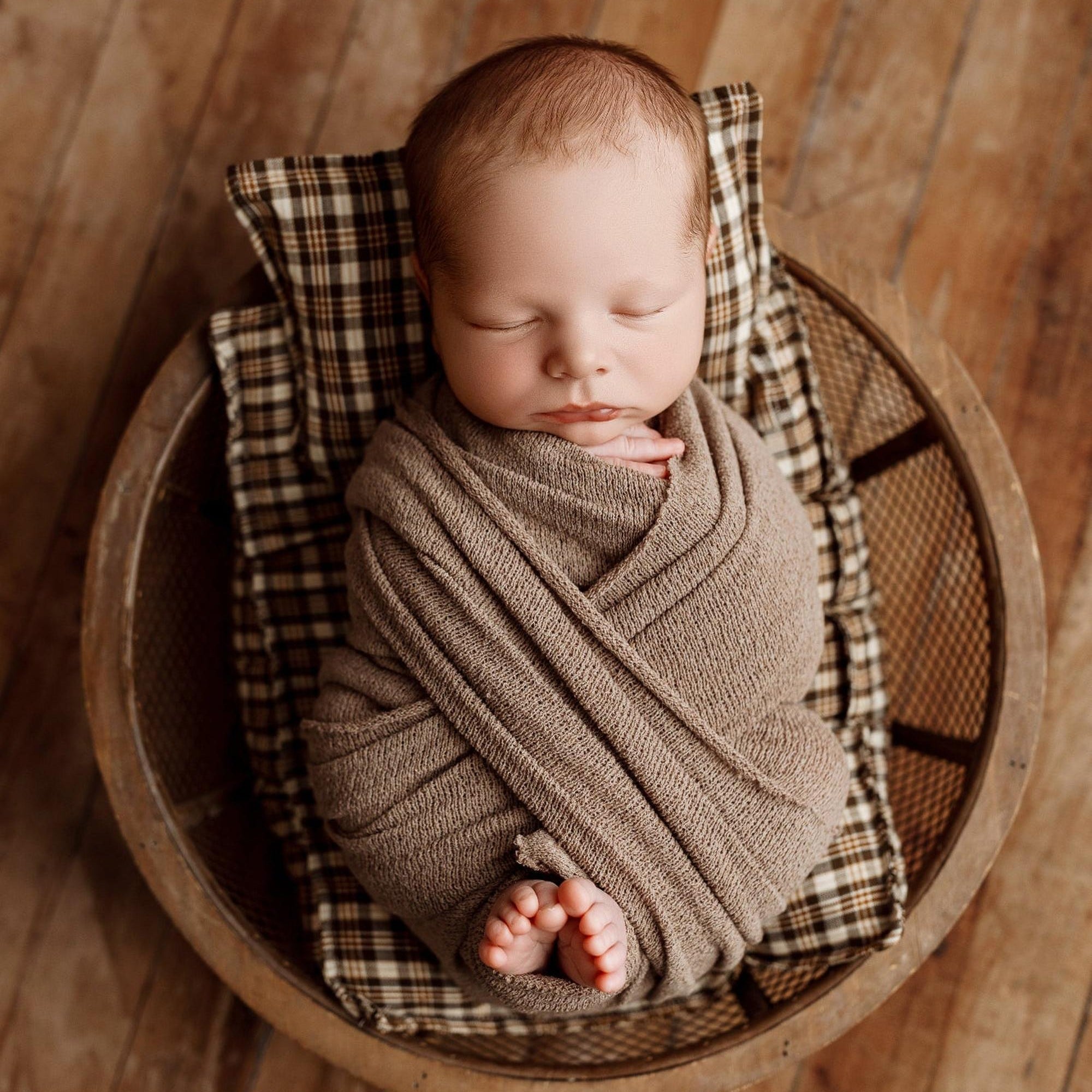 Newborn baby wrapped in a coffee brown knit blanket, resting on brown plaid sheets and a matching pillow in a round basket.
