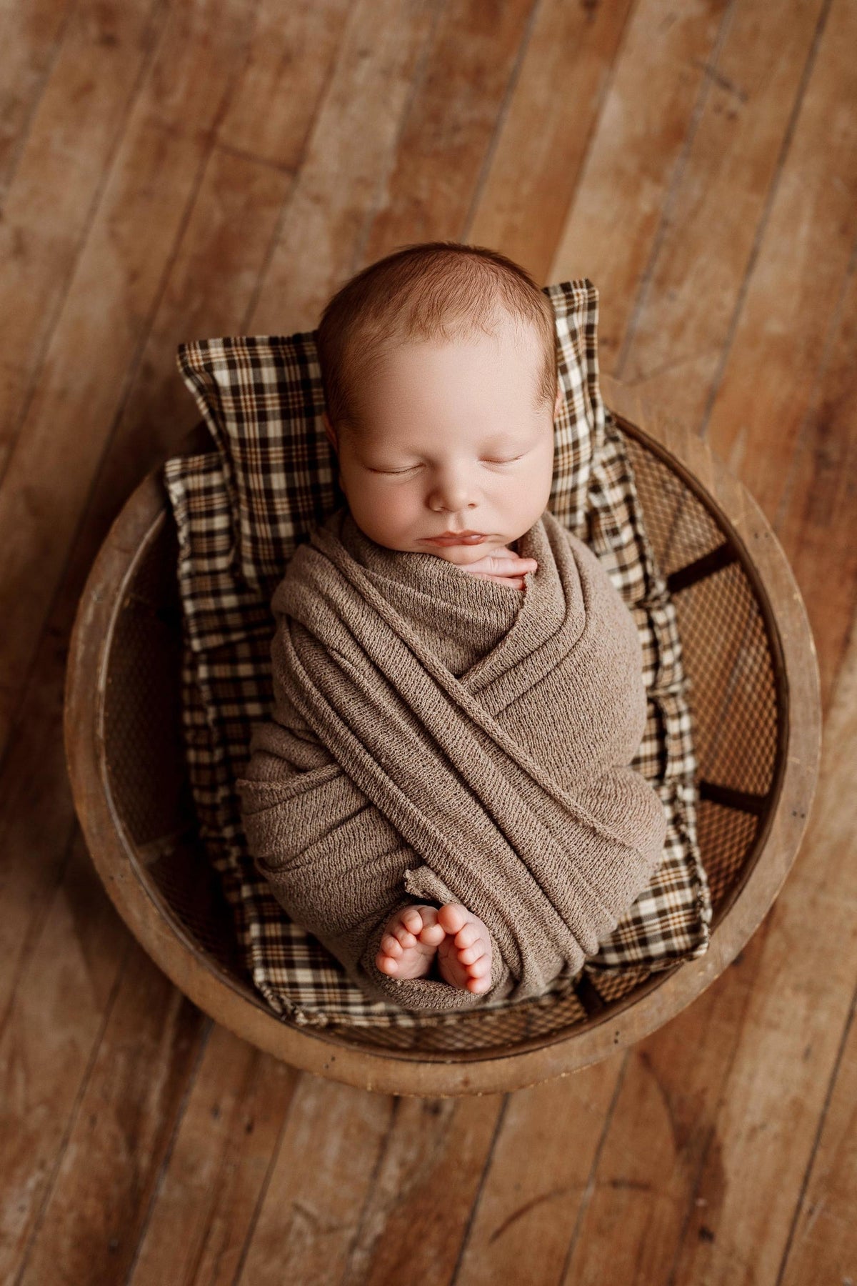 Newborn baby wrapped in a coffee brown knit blanket, resting on brown plaid sheets and a matching pillow in a round basket.