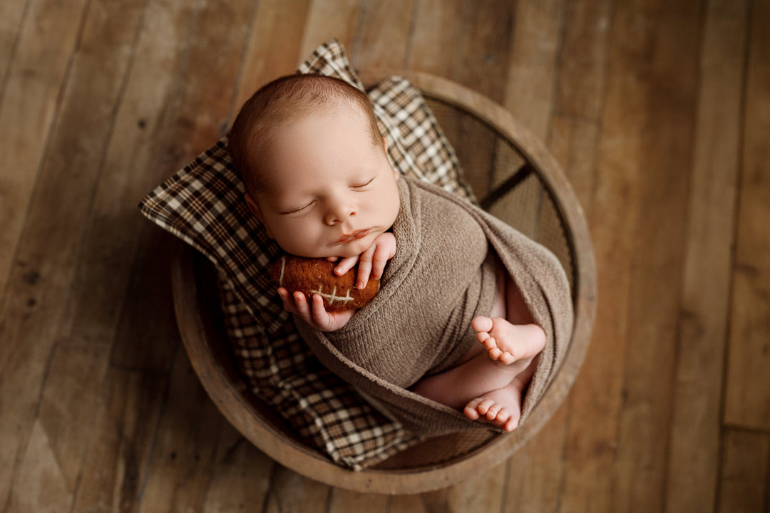 Newborn wrapped in the Nest | Coffee Bean wrap, holding a small felt football while posed on brown plaid pillow and sheet set.