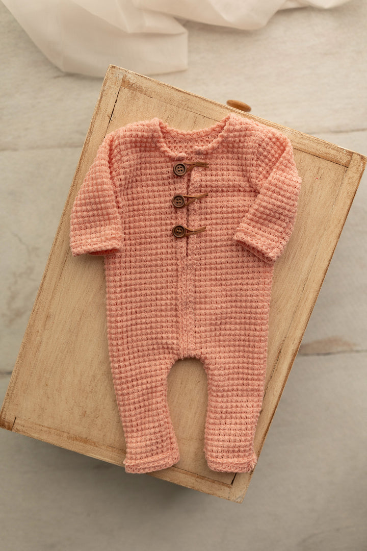 Peach knit newborn romper with wooden button details, styled flat on a wooden crate for photography use.