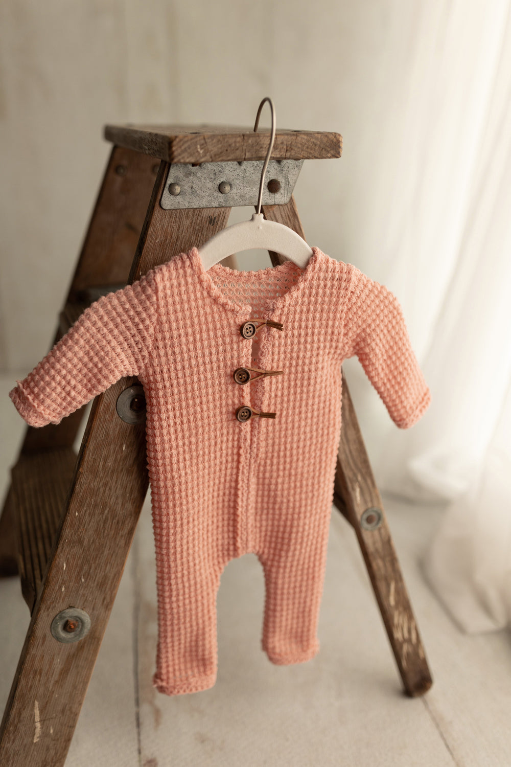 Pink knit newborn romper with three wooden buttons, displayed hanging on a ladder for photography styling.