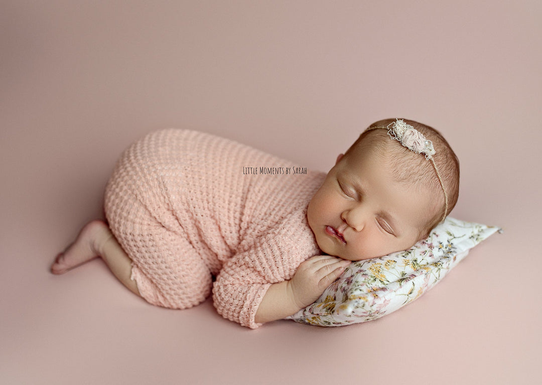 Newborn wearing a pink knit romper with wooden buttons, posed on a floral pillow during a photography session.