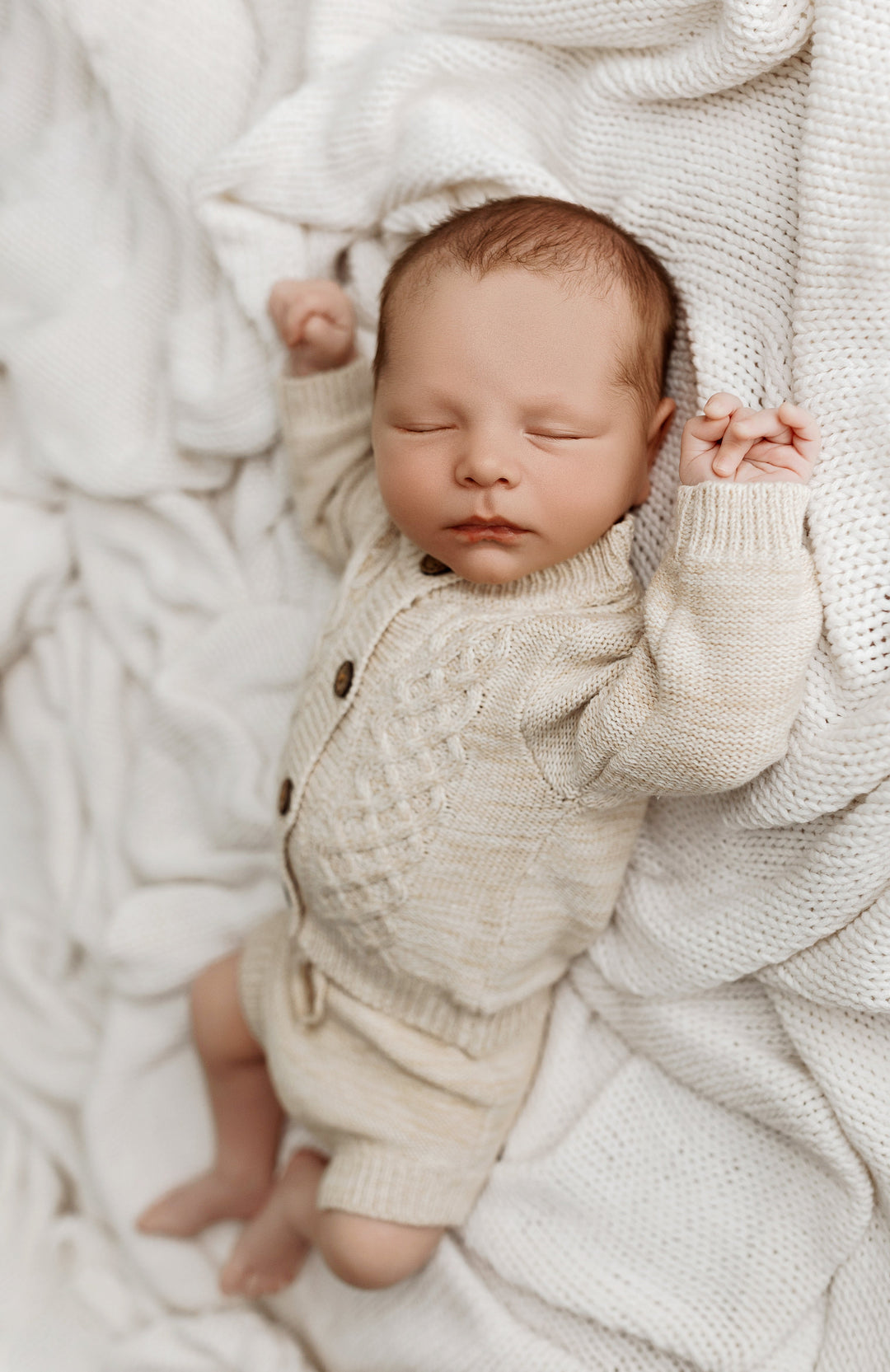 Newborn posed in the Woodland knit set with cable-knit button cardigan and matching shorts, laying on a textured cream blanket for photography.